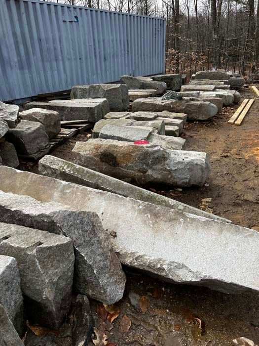 A large blue shipping container stands near a pile of assorted gray stone blocks and slabs in a dirt-covered yard.