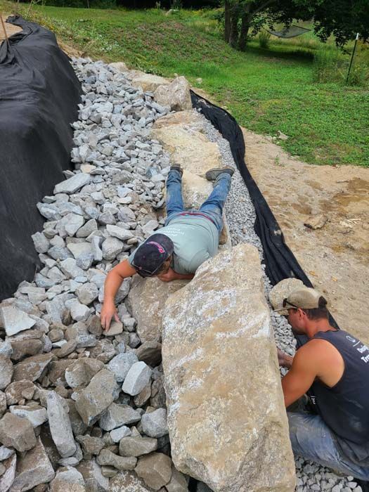 Two workers place large rocks and gravel along a sloped hillside lined with black fabric for erosion control.