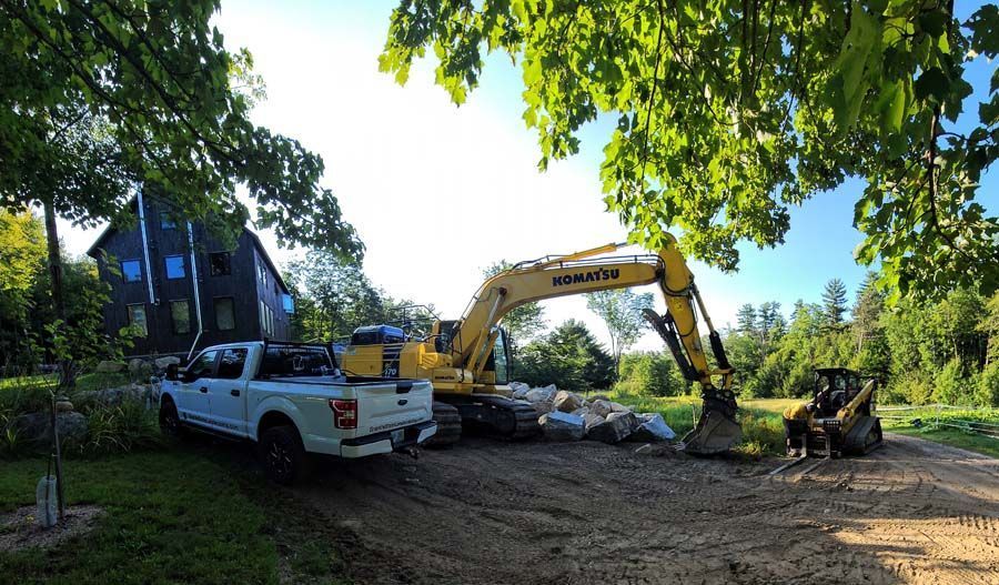 A yellow Komatsu excavator, a white pickup truck, and a skid steer parked on a dirt lot near a house and trees.