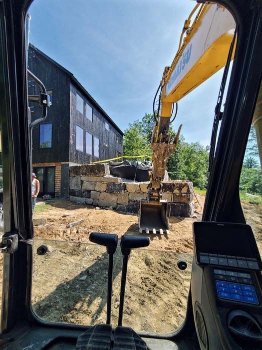 View from inside an excavator cab looking toward a stone retaining wall being built near a modern black house.
