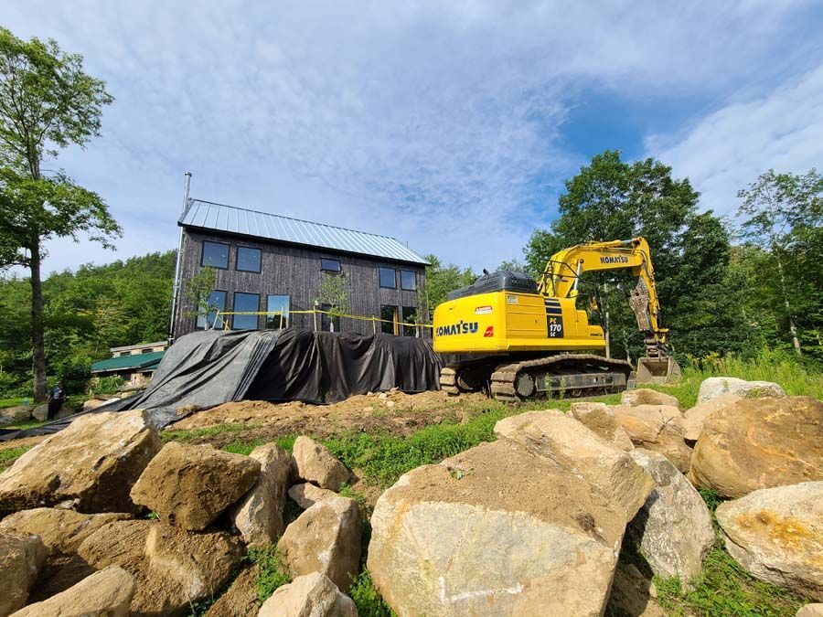 A yellow excavator sits in front of a modern, dark-wood building under construction, surrounded by large rocks and greenery.