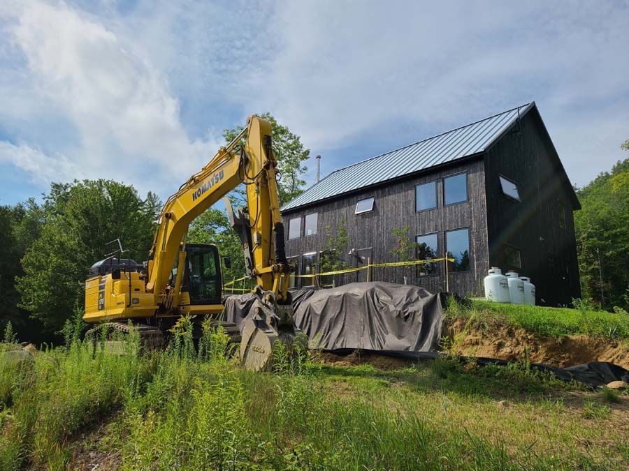 A yellow excavator sits in front of a modern, dark-sided building near a grassy field under a cloudy blue sky.