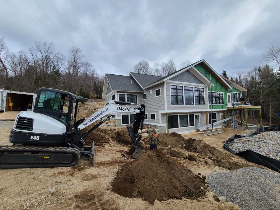 A white excavator digs a trench in front of a modern house under construction on a cloudy day.