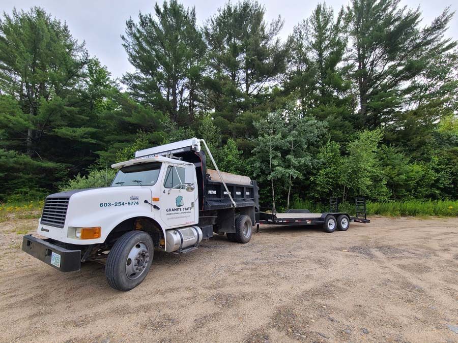 A white dump truck with a black trailer attached, parked on a gravel lot in front of a dense line of green trees.