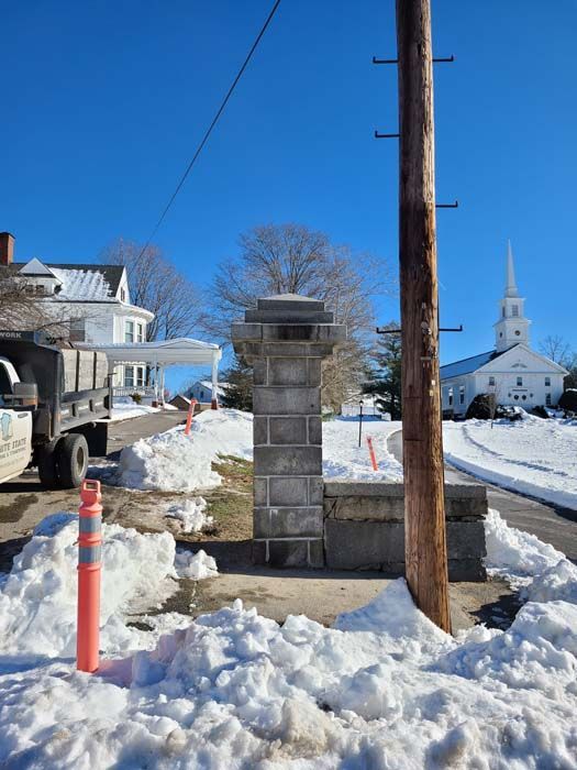 A snow-covered road scene features a stone pillar, a utility pole, a dump truck, and a white church steeple in the distance.