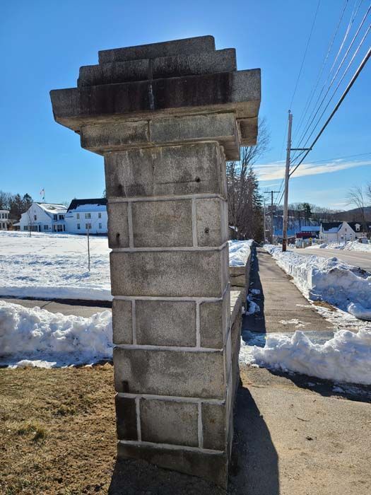 A stone masonry gate pillar stands on a snowy sidewalk near a road on a sunny day.