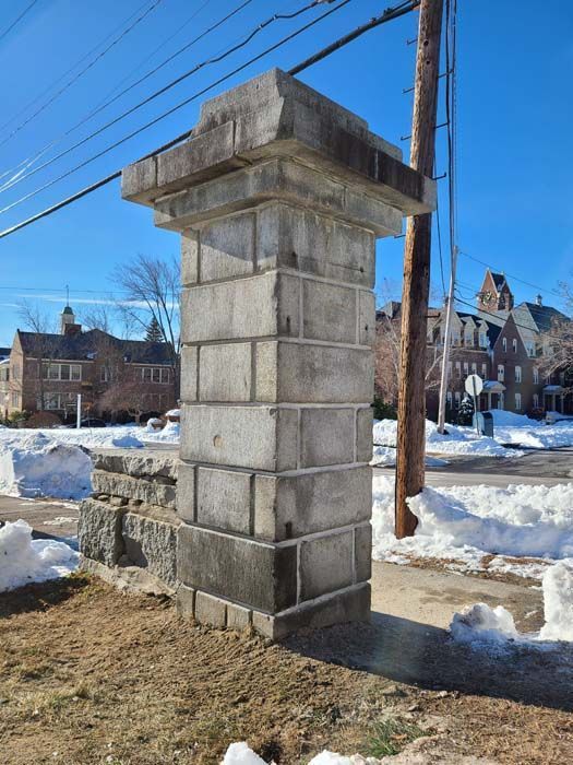 A tall stone pillar stands on a snowy roadside with large historic buildings in the background under a clear blue sky.