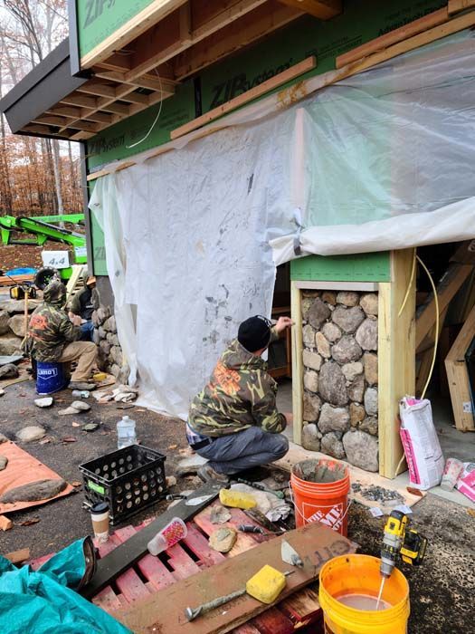 Two workers in camouflage jackets apply stones to a residential foundation wall at an active outdoor construction site.
