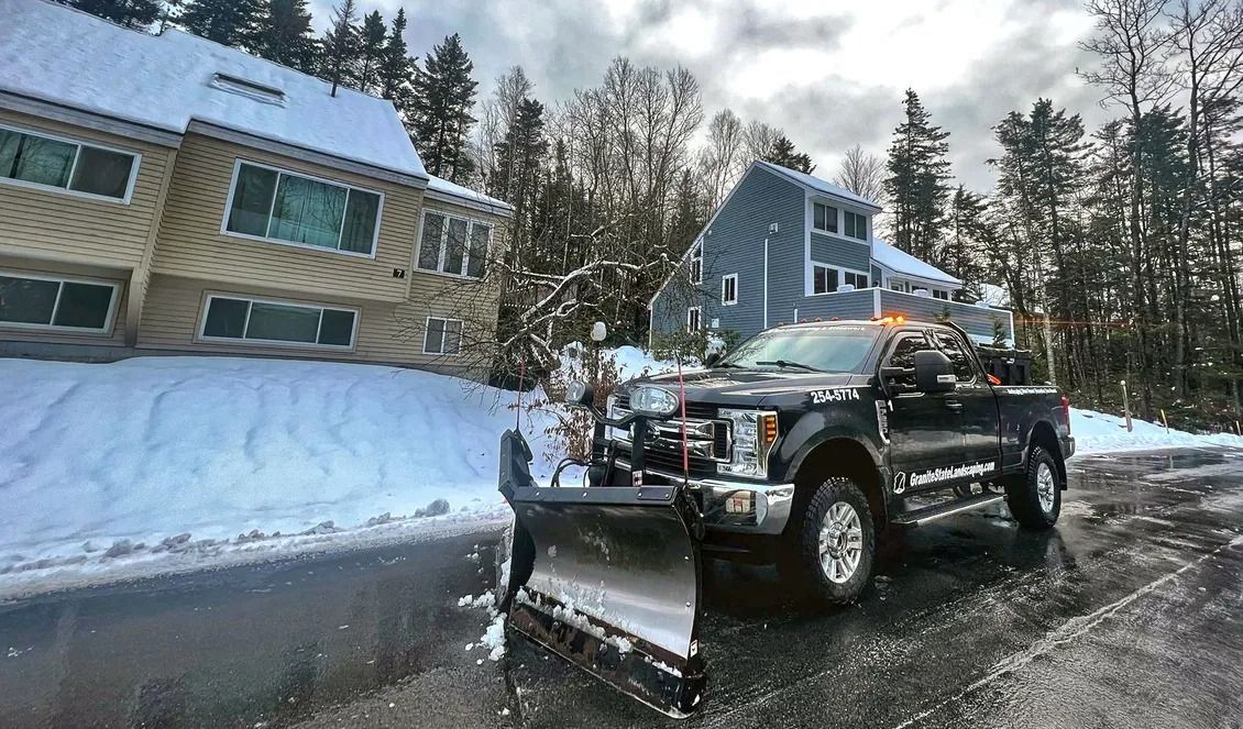 A black pickup truck with a snow plow attached parked on a snowy road in front of residential houses.