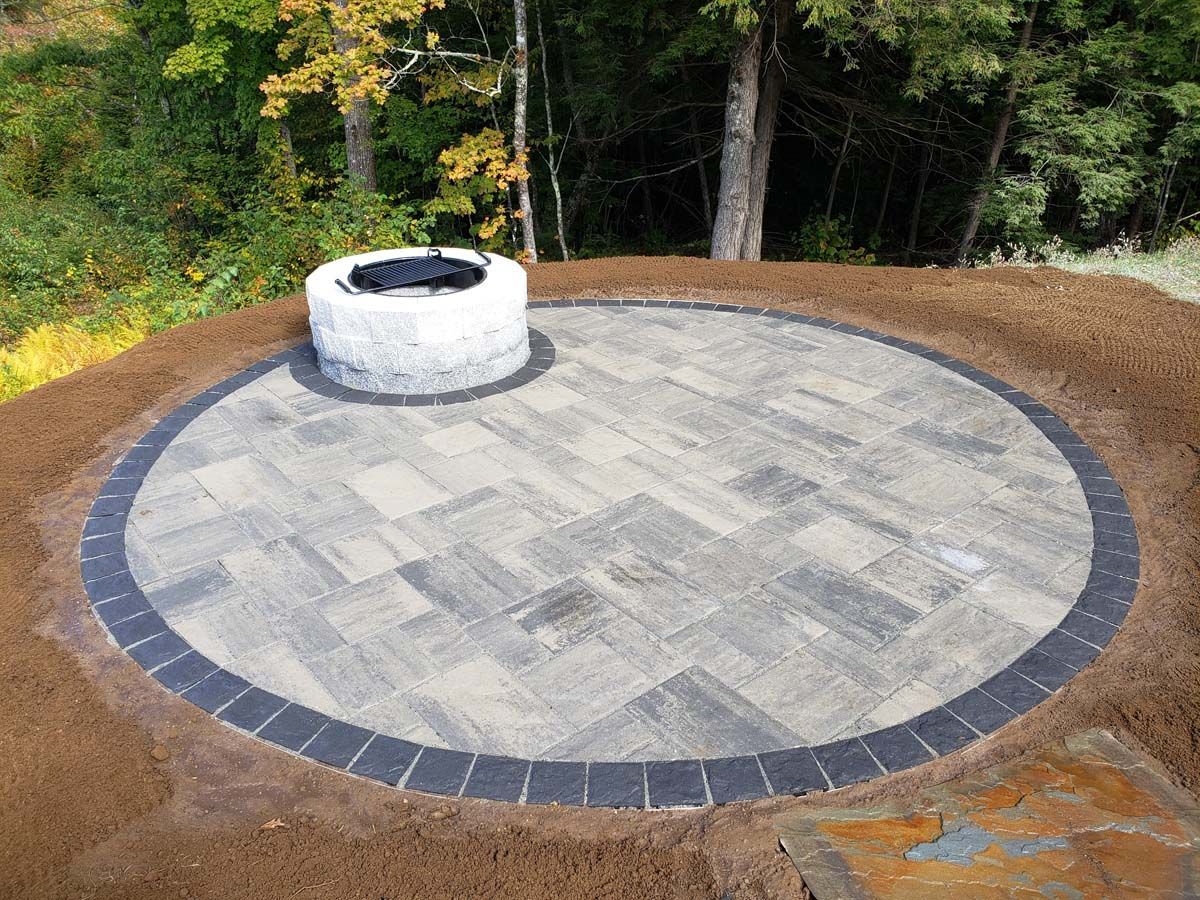 A circular stone paver patio with a matching circular fire pit, set in a backyard area with trees in the background.