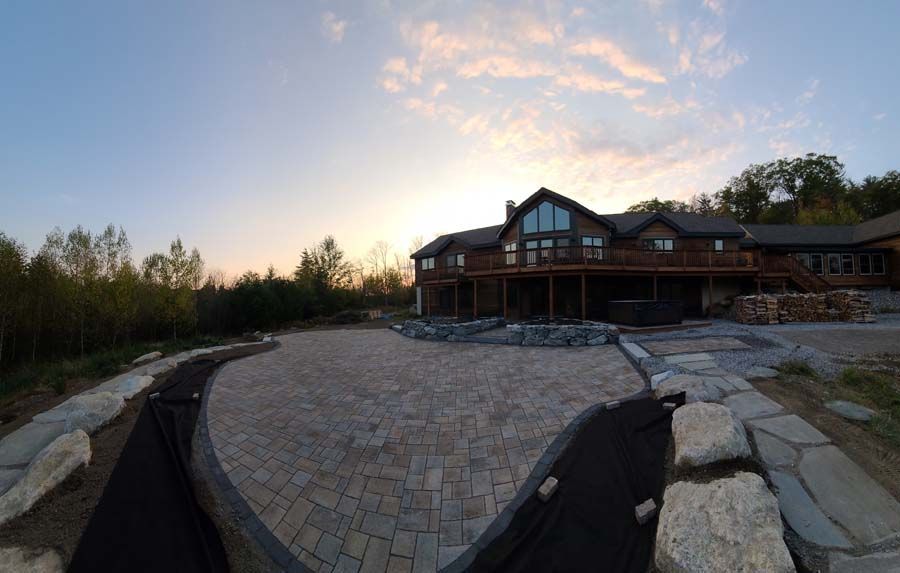 A large wooden house with a paved stone patio under a sunset sky, surrounded by trees and decorative boulders.