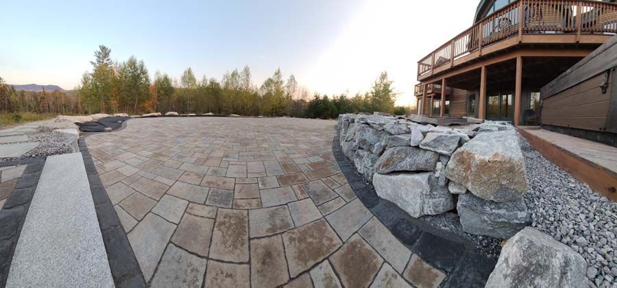A wide-angle view of a backyard patio with stone pavers, a natural stone retaining wall, and a wooden deck above.