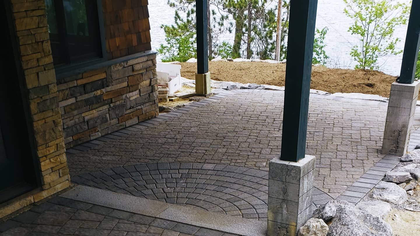 A stone-paved patio with a curved pattern under a covered porch overlooking trees and water.