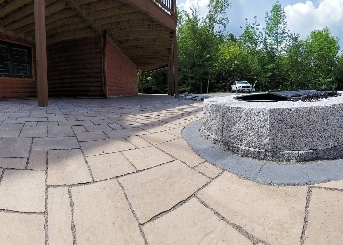 A stone fire pit on a paver patio beneath a wooden deck, with a wooded backdrop and a white vehicle parked in the distance.