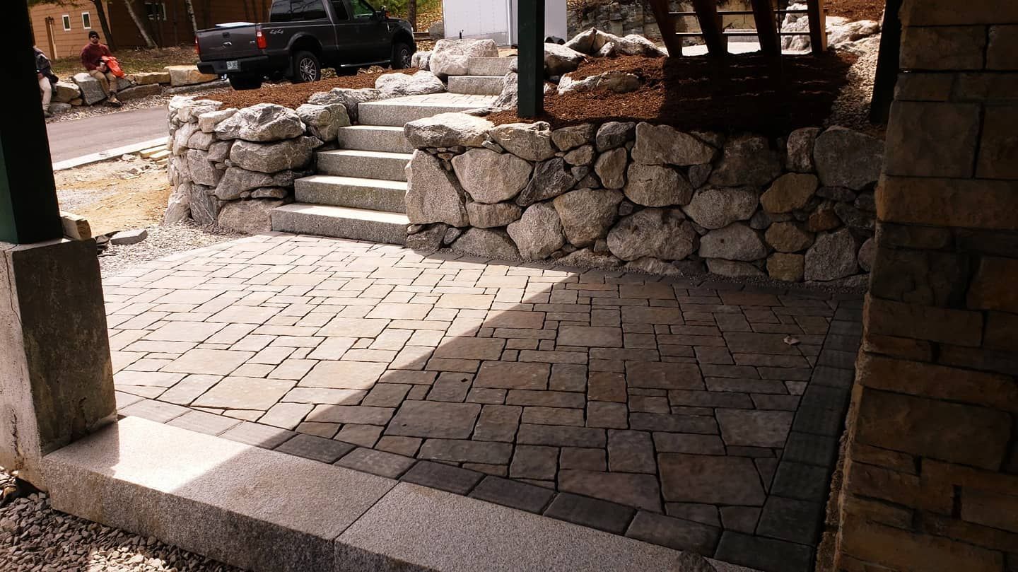 A stone patio features two patterns of pavers leading to stone steps and a retaining wall in a partially shaded outdoor area.