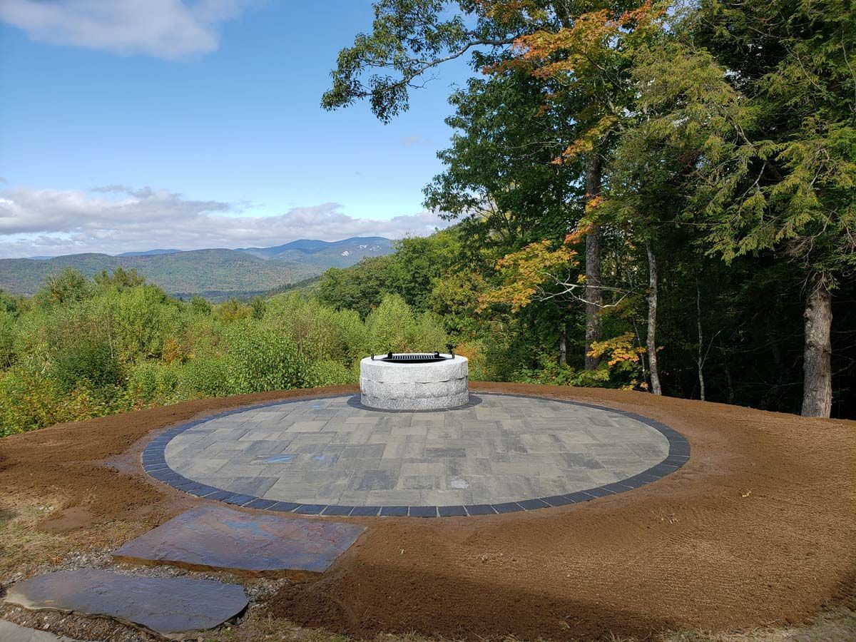 A circular stone patio with a fire pit overlooking a mountainous landscape, surrounded by trees and mulch.