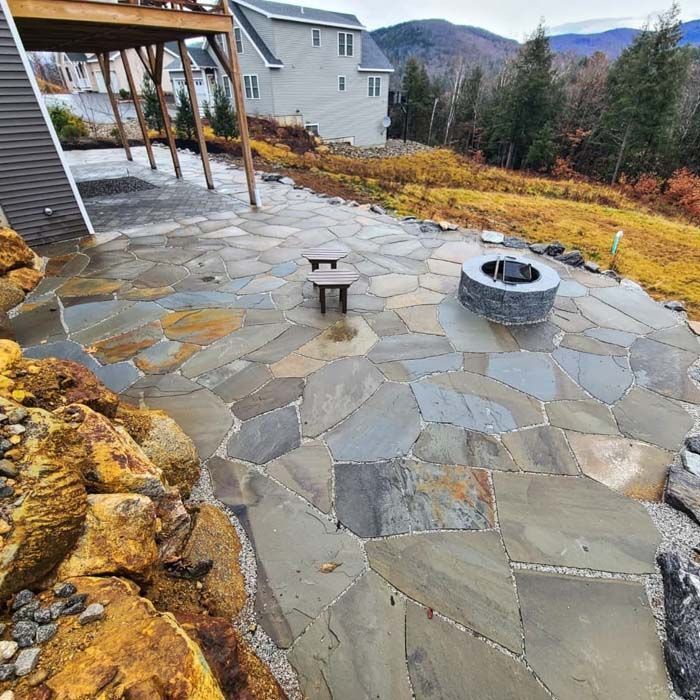 A flagstone patio featuring a circular stone fire pit, set beside a house with a wooden deck overlooking distant mountains.