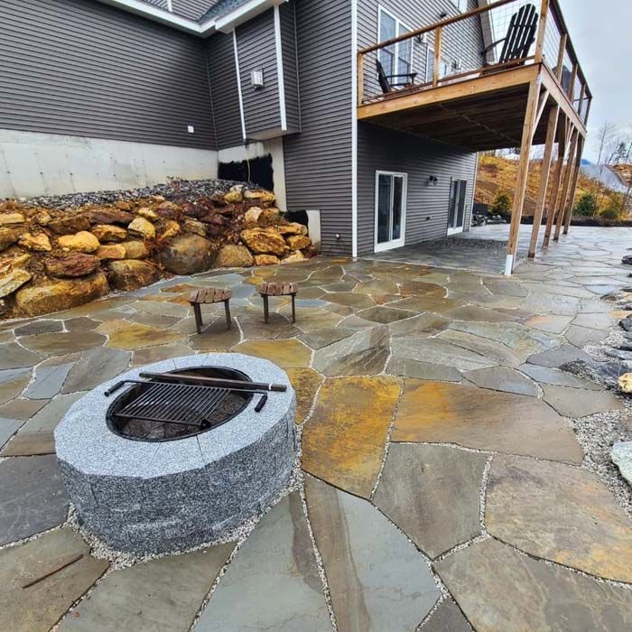 A gray stone fire pit sits on a flagstone patio beside a house with a wooden deck and a stacked rock retaining wall.