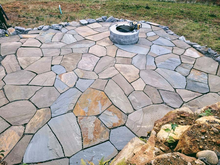 Flagstone patio with a circular stone fire pit, set in a natural, grassy landscape with rock border elements.