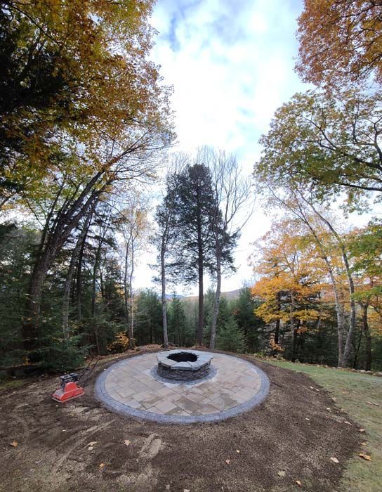 A circular stone fire pit sits in the center of a newly installed stone patio, surrounded by trees with autumn foliage.