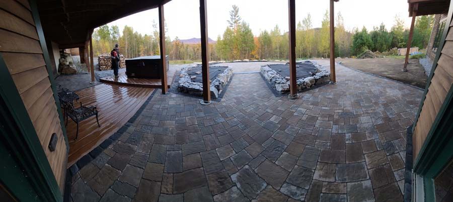 A wide patio with stone pavers, a wooden deck, a hot tub, and support posts looking out toward a forest and mountains.