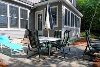 A patio set with a round table, umbrella, and chairs, next to a blue lounge chair on a paved patio beside a gray house.