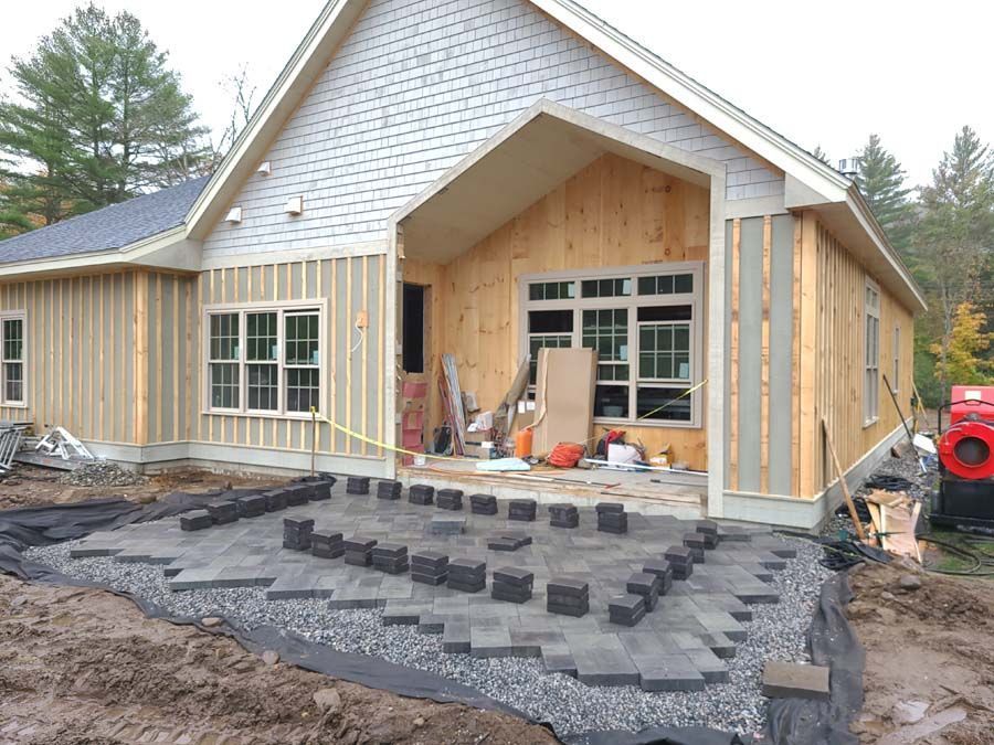 Construction site featuring a house with wood-paneled walls and a partially paved stone patio with stacks of loose pavers.