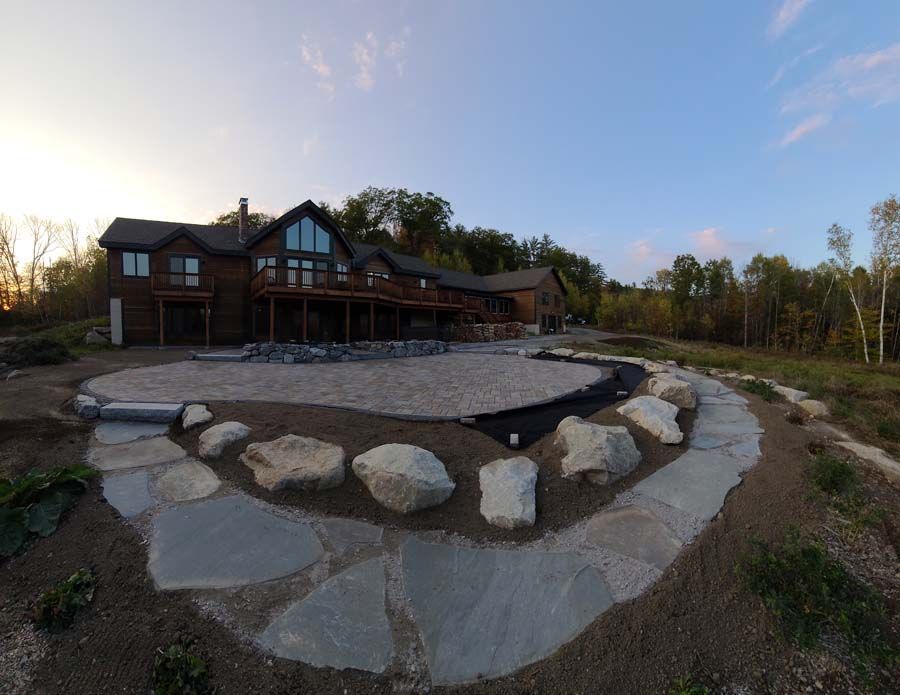 A multi-story wooden house with a large stone patio under construction, bordered by large rocks and a stone walkway.