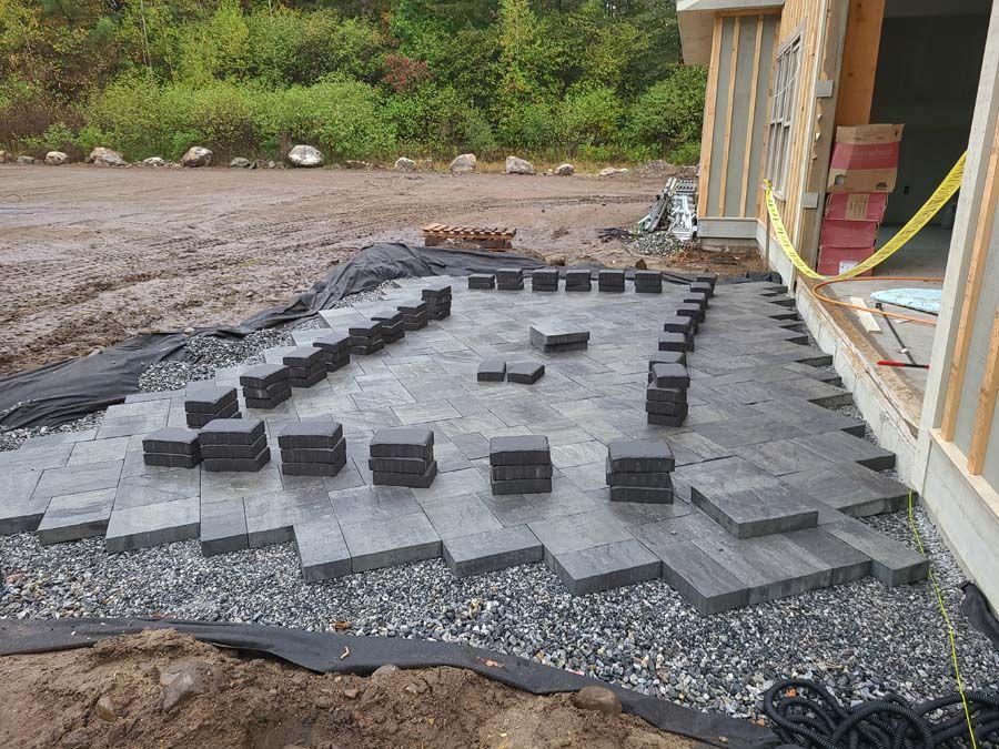 Gray paving stones being laid in a geometric pattern on a gravel base next to an unfinished building under construction.