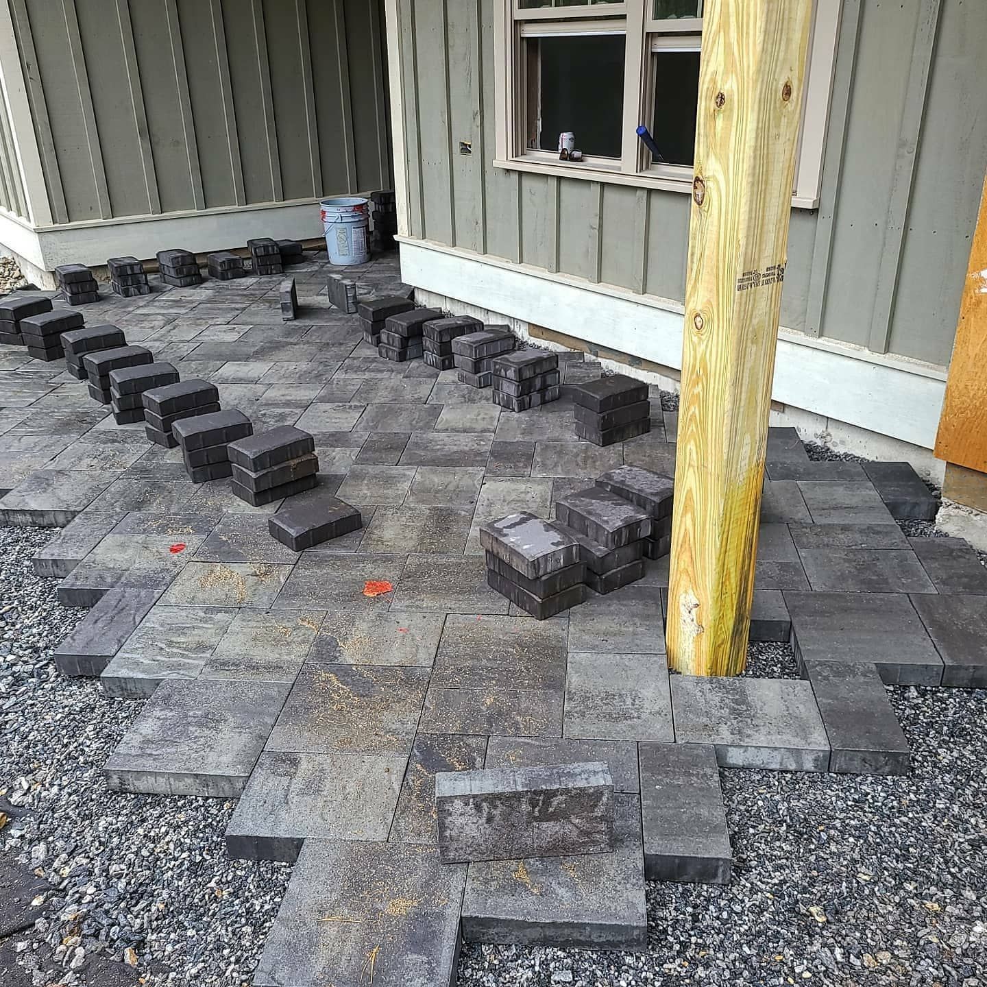 A partially completed patio installation with dark stone pavers laid in a pattern around a wooden porch support post.