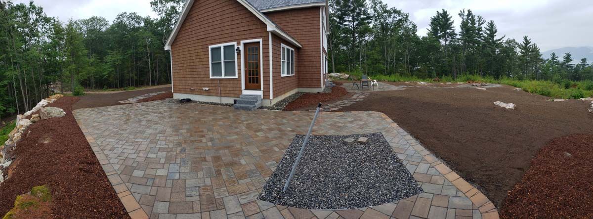 A brown house with a new brick paver patio featuring a central gravel square, surrounded by mulch beds and forest trees.