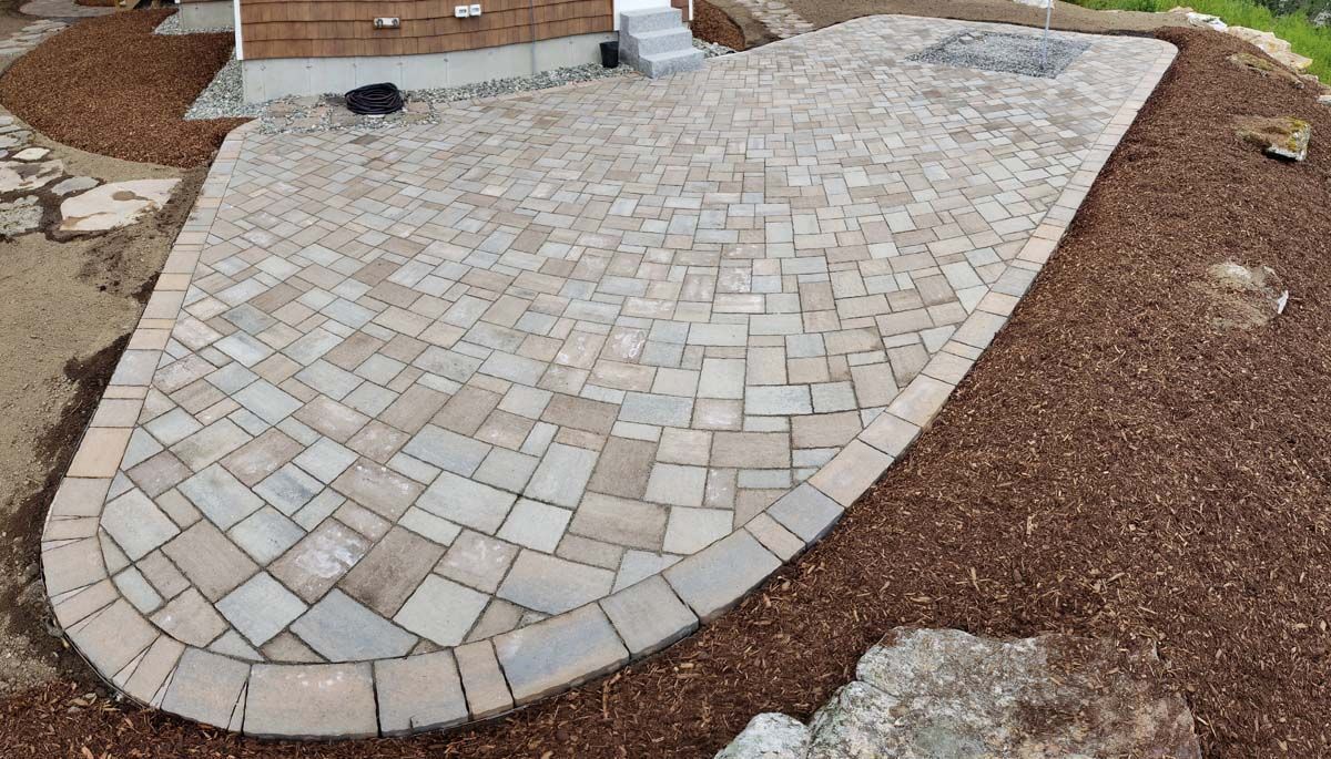 A light-gray paver patio with a curved border, surrounded by brown mulch in a residential yard.
