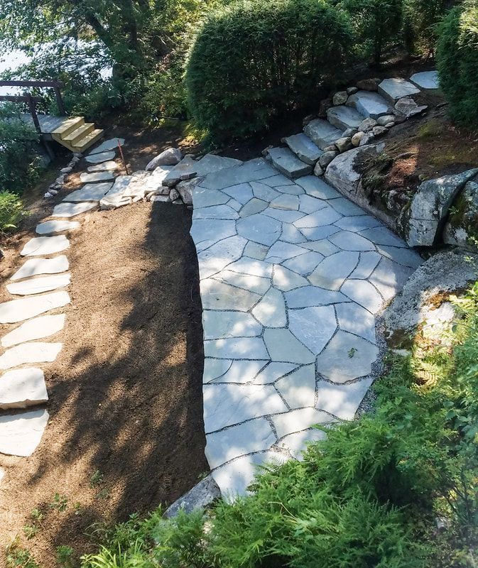 A stone-paved patio and walkway in a garden setting, with natural stone steps ascending beside a dark soil area.