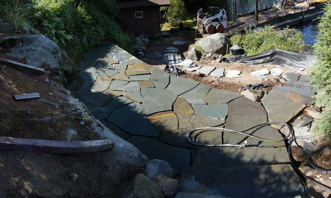 A stone patio under construction with irregular slate pieces set in mortar, nestled in a wooded outdoor area.