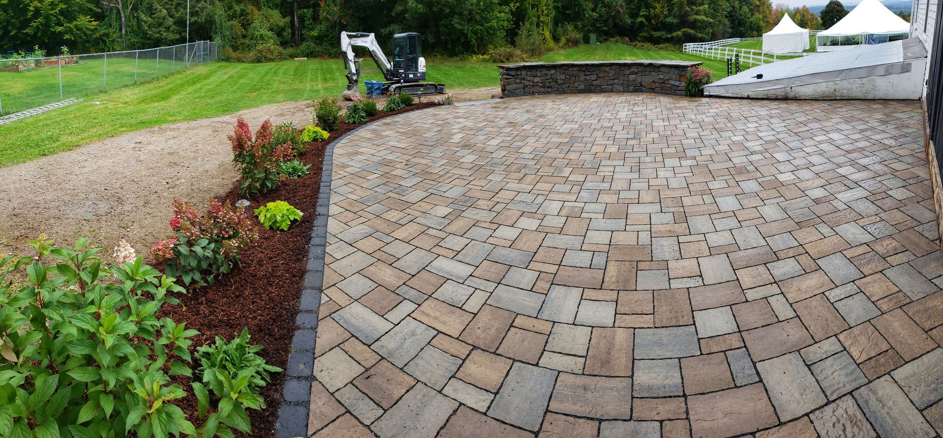 A newly installed stone paver patio sits adjacent to a landscaped garden bed with mulch and shrubs in a grassy yard.