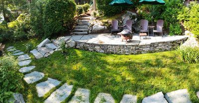 A stone path winds through a green lawn toward a raised patio with chairs, a fire pit, and two umbrellas in a garden.