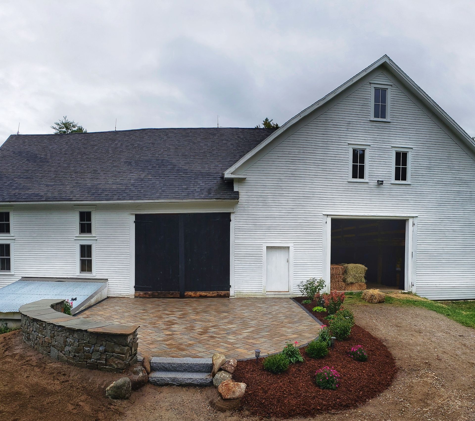 A white, horizontal-log barn with a black door and a stone patio featuring a curved retaining wall and a mulched garden.
