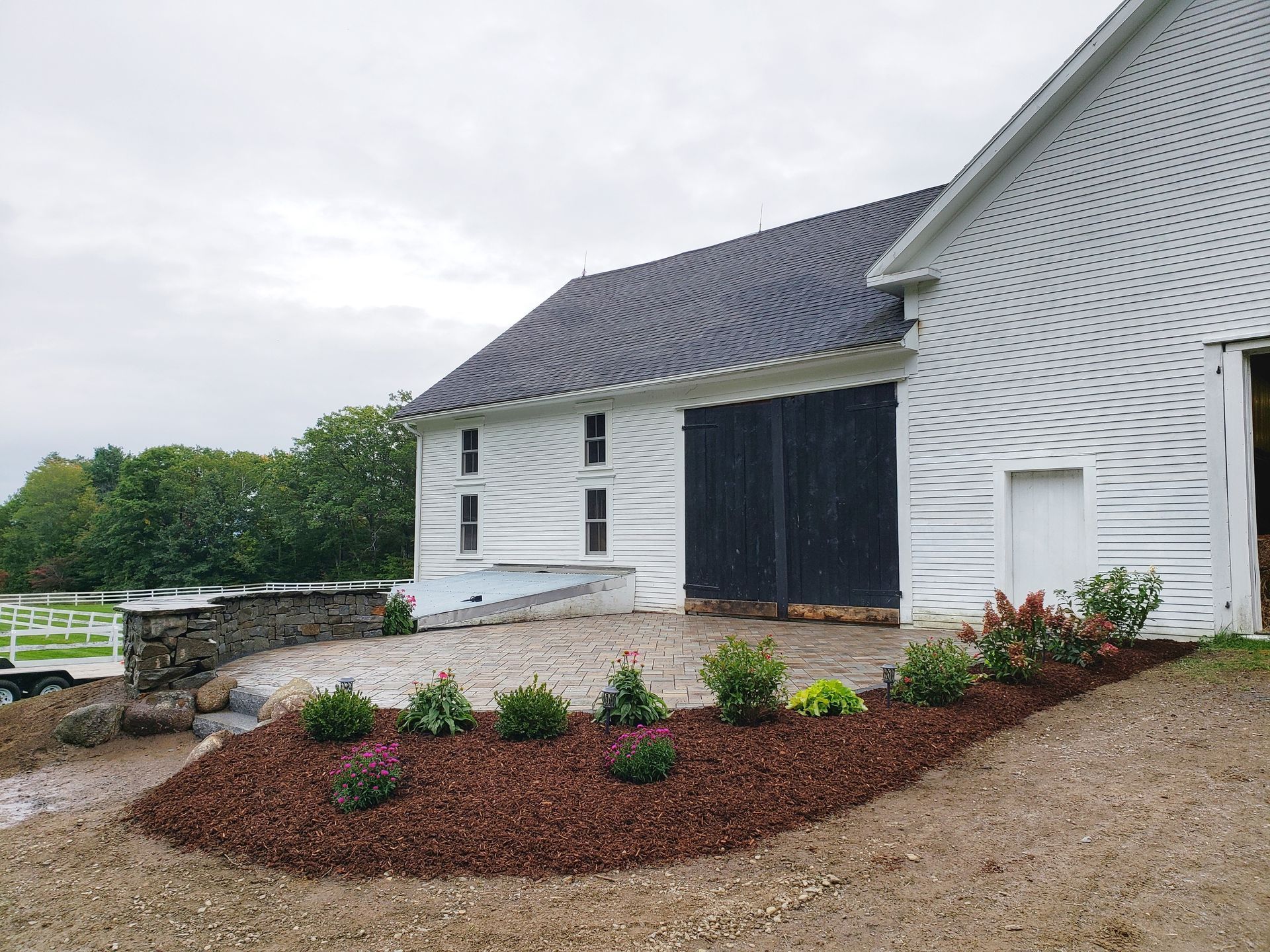 White barn with a gray roof and black doors next to a stone patio with mulched landscaping and shrubs.