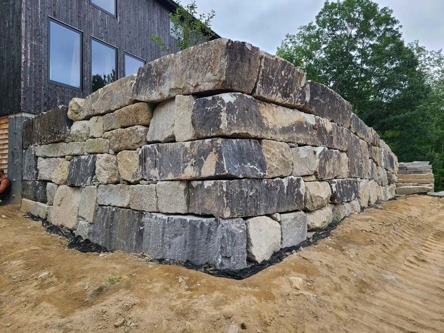 A large, stacked stone retaining wall at the corner of a dark, modern house with a dirt yard in the foreground.
