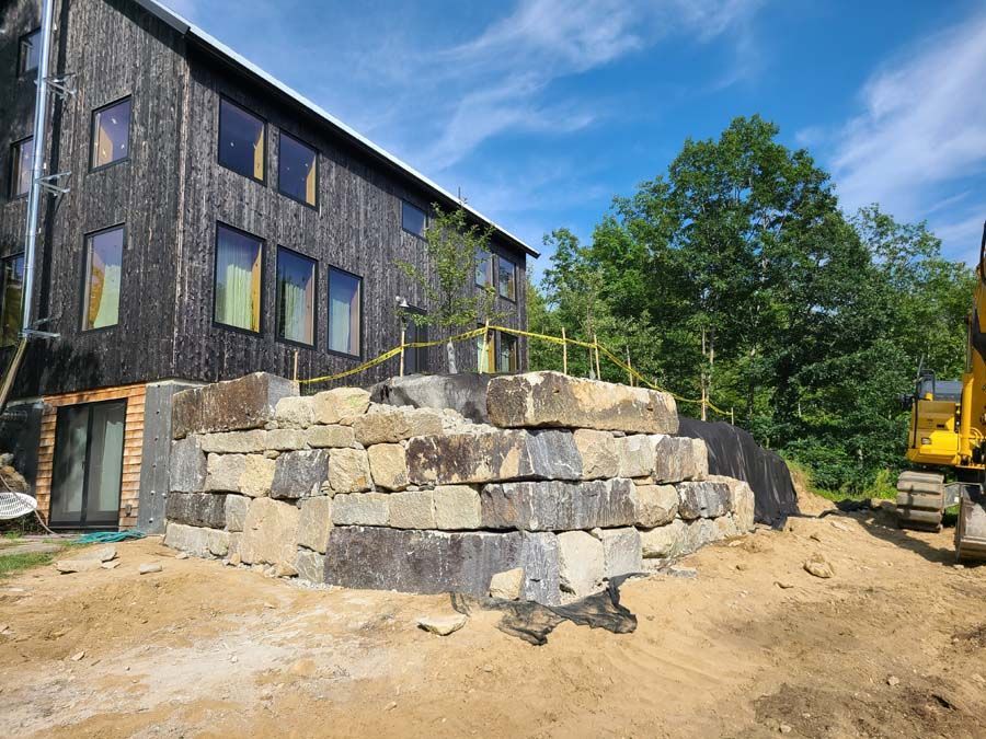 A large stone retaining wall stands in front of a dark-sided building at a construction site with an excavator nearby.