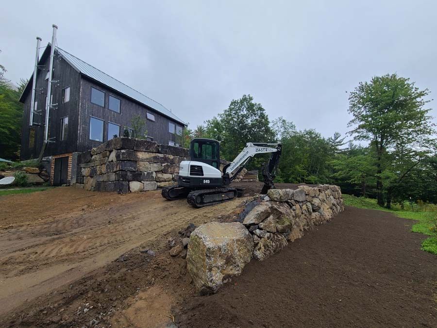 A white mini excavator sits on a dirt construction site next to a large rock retaining wall and a dark, modern home.