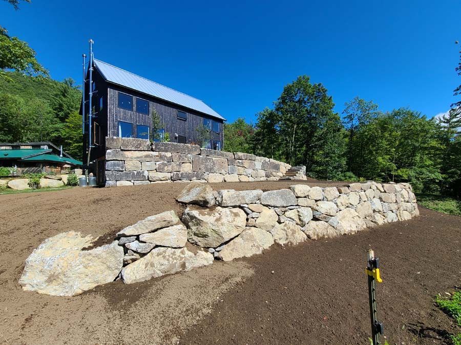 A dark, modern house stands atop a tiered stone retaining wall set against a lush forest and clear blue sky.