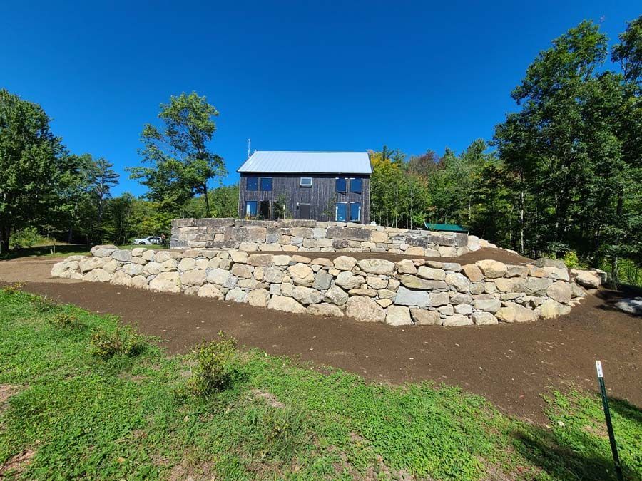 A modern, dark-sided house with a metal roof sits atop a large, multi-tiered natural stone retaining wall on a sunny day.