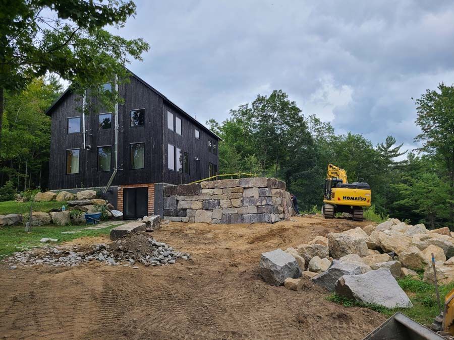 A dark wood house sits near a large stone retaining wall under construction, with a yellow excavator nearby.