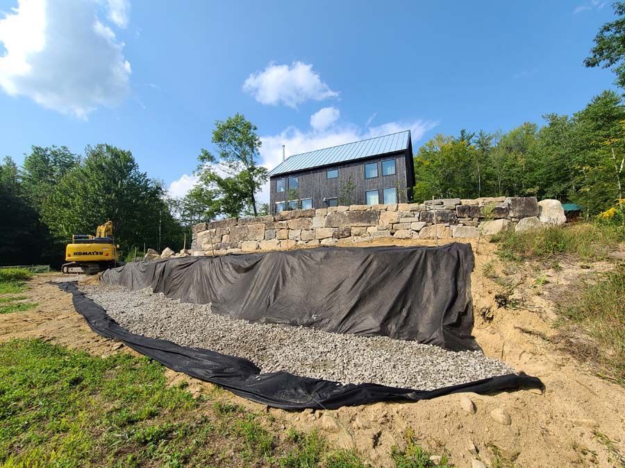 A modern house sits behind a tiered landscaping project featuring a stone wall, black geotextile fabric, and gravel fill.