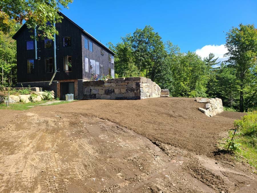 A black house sits on a hill above a freshly leveled dirt yard with a large stone retaining wall.