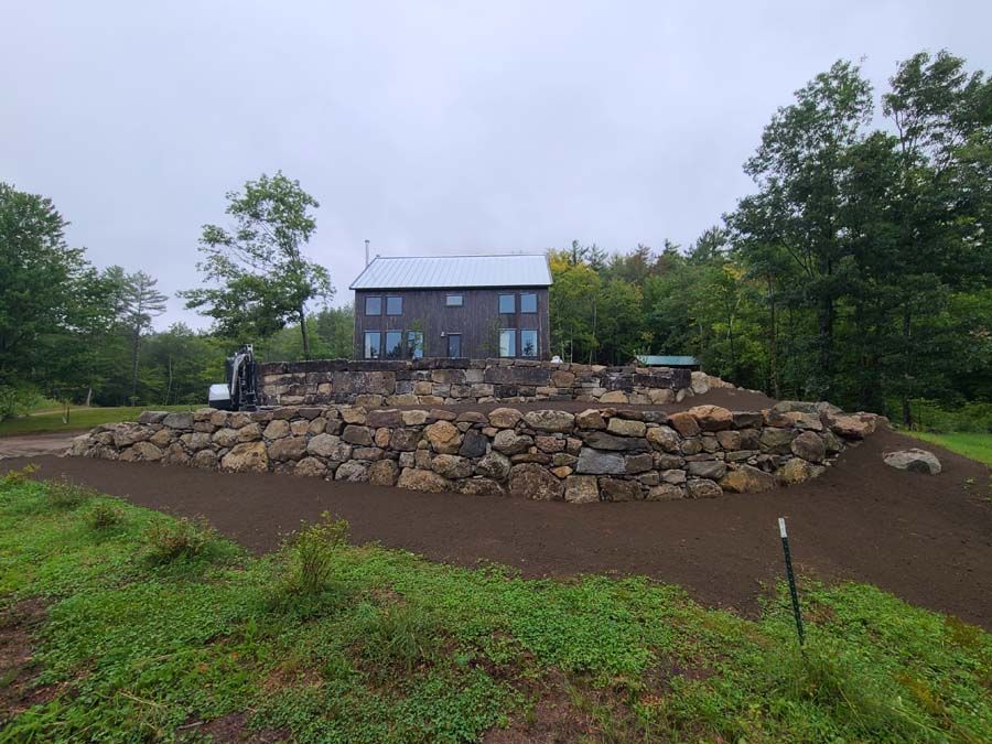A dark, modern house sits atop a tiered stone retaining wall, surrounded by green trees and fresh, dark soil.