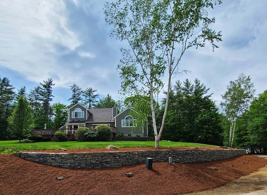 A house with grey siding sits behind a natural stone retaining wall and a sloped lawn surrounded by lush green trees.