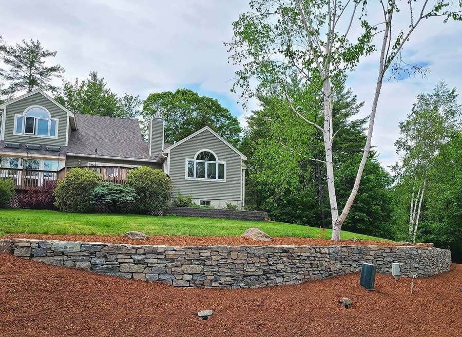 A stone retaining wall curves across a lawn in front of a gray-sided suburban house with white trim and a birch tree.