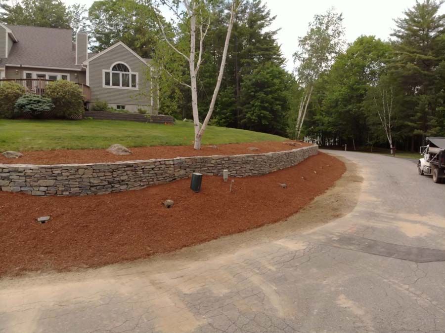 A stone retaining wall curves along the edge of a grassy yard, bordered by brown mulch and a paved driveway.